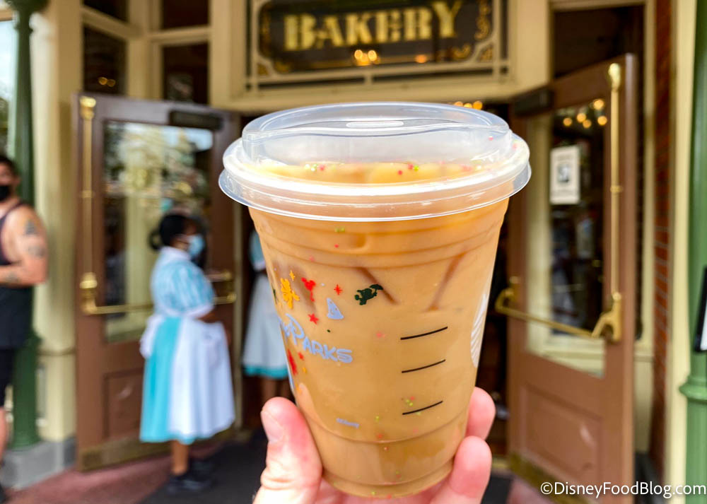 https://www.disneyfoodblog.com/wp-content/uploads/2021/11/2021-wdw-disney-world-magic-kingdom-main-street-bakery-iced-sugar-cookie-latte-with-almond-milk-holiday-drinks-in-front-of-bakery.jpg