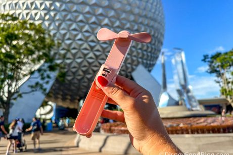 A handheld JISULIFE fan, a portable personal cooling device, is shown in the foreground with the iconic Spaceship Earth geodesic sphere at Epcot in Walt Disney World in the background.
